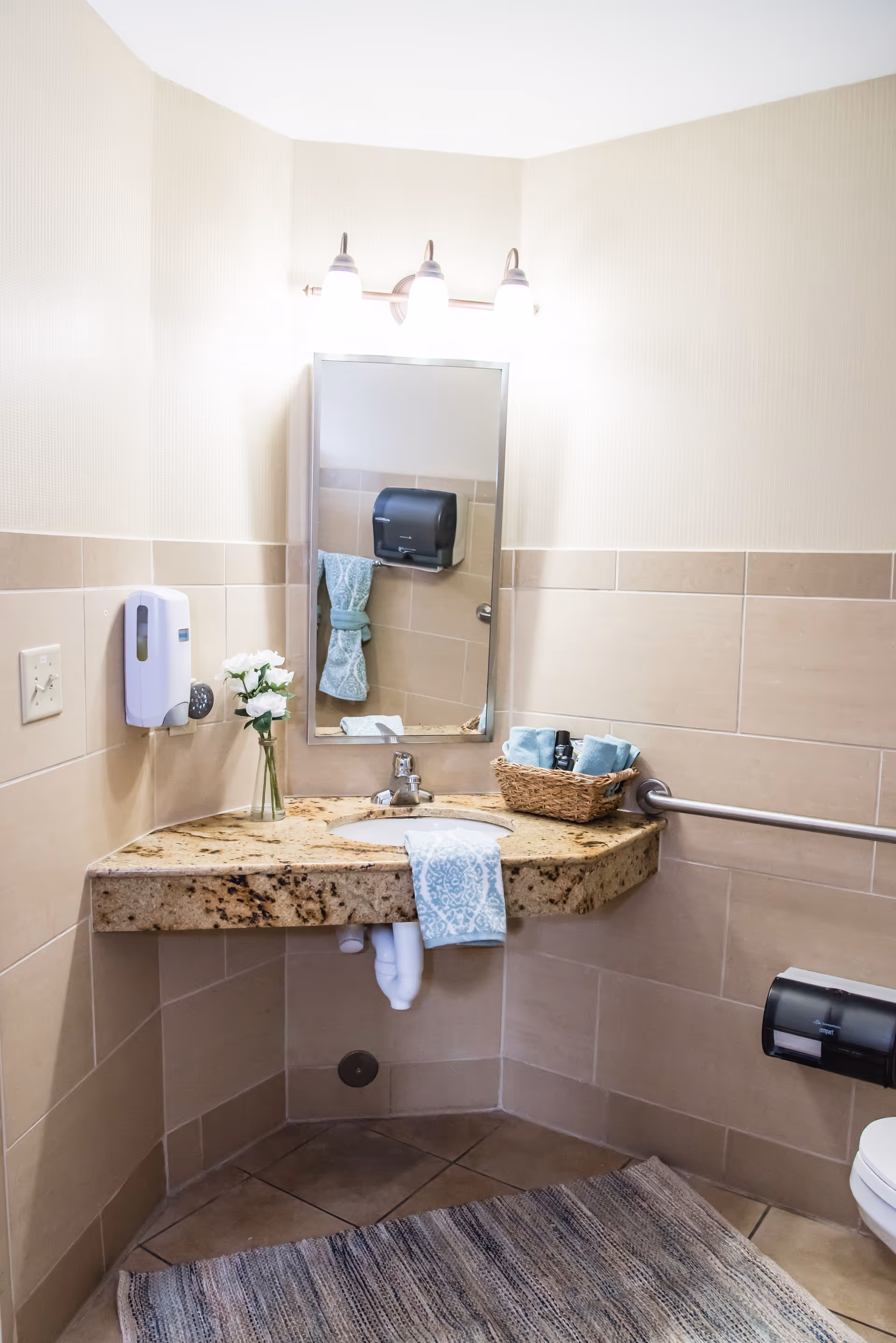 A clean and well-lit bathroom corner with a granite countertop sink, a rectangular mirror above it, and three light fixtures mounted on the wall. There is a small vase with white flowers on the left side of the countertop and a basket with rolled blue towels and toiletries on the right. A blue patterned hand towel hangs over the front edge of the countertop. The walls are tiled halfway up with beige tiles, and there is a handrail on the right side. A soap dispenser is mounted on the left wall, and a paper towel dispenser is visible on the right wall near the toilet. A textured rug covers the tiled floor.