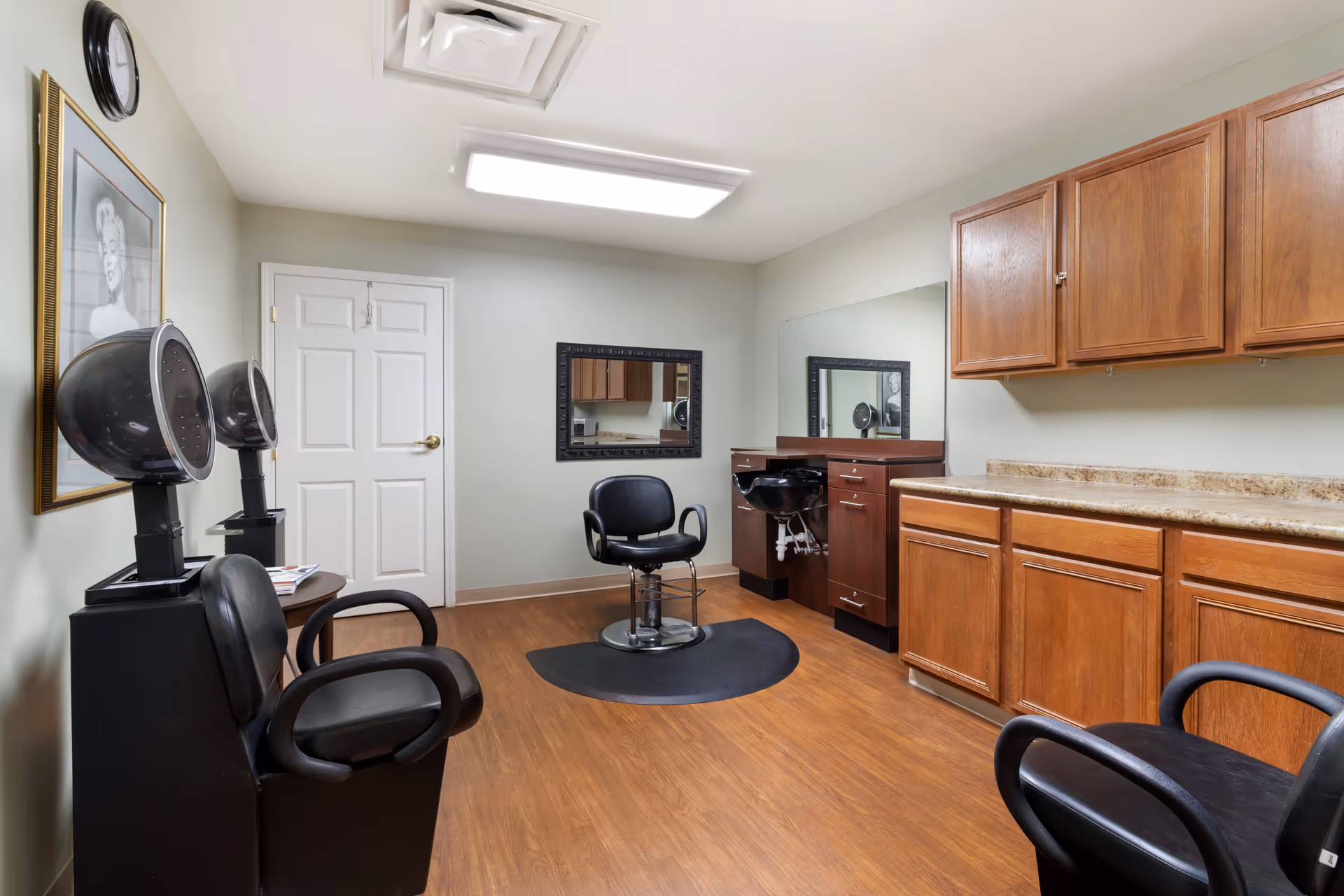 Interior view of a hair salon room in a senior living facility with wooden flooring, a black salon chair in front of a mirror, two black hair dryers, wooden cabinets, and a framed black and white portrait on the wall.