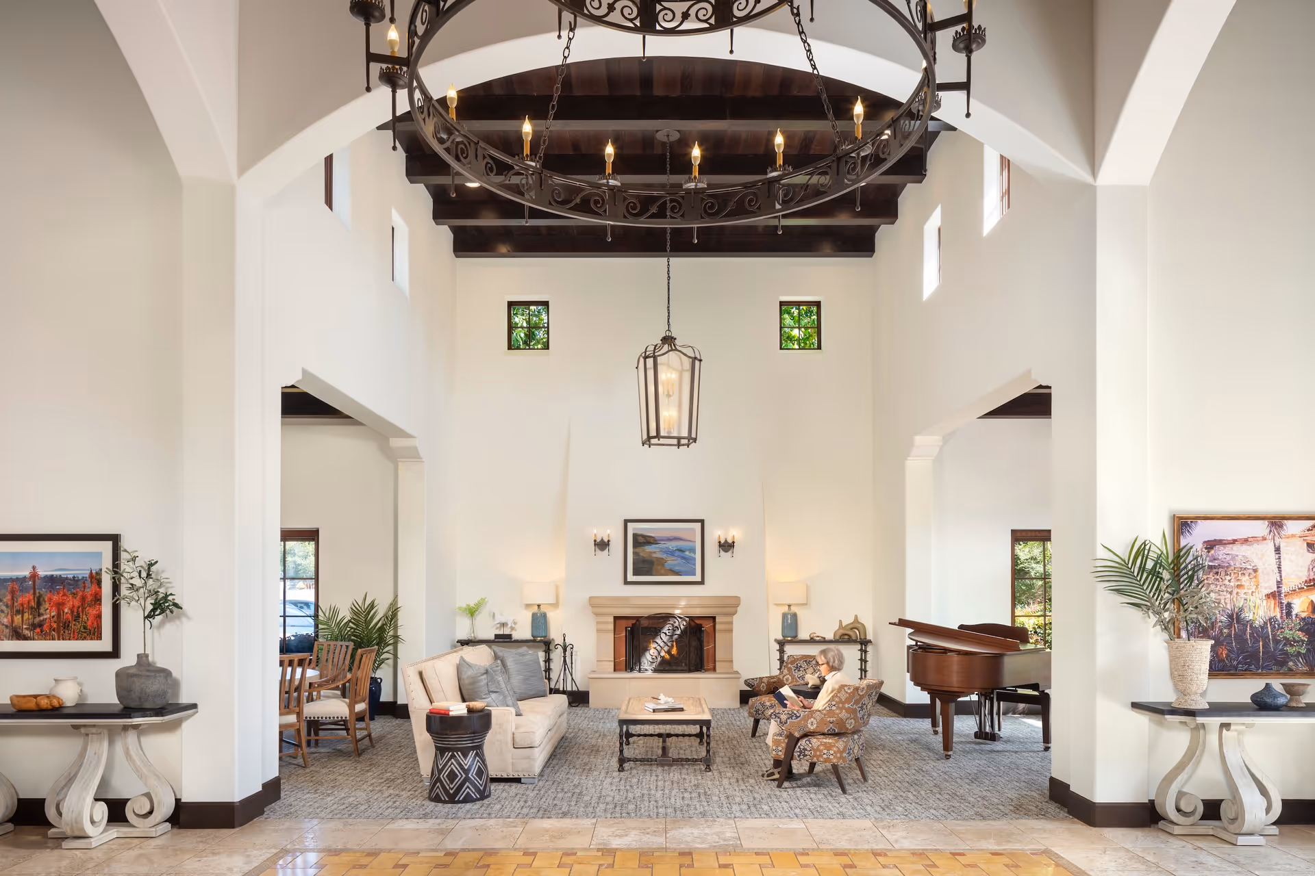A spacious and elegant living room area with high ceilings and exposed wooden beams. The room features a large chandelier, a fireplace with a framed picture above it, a beige sofa, patterned armchairs, a coffee table, and a grand piano. There are decorative plants and framed artwork on the walls. A person is seated in one of the armchairs reading a book.