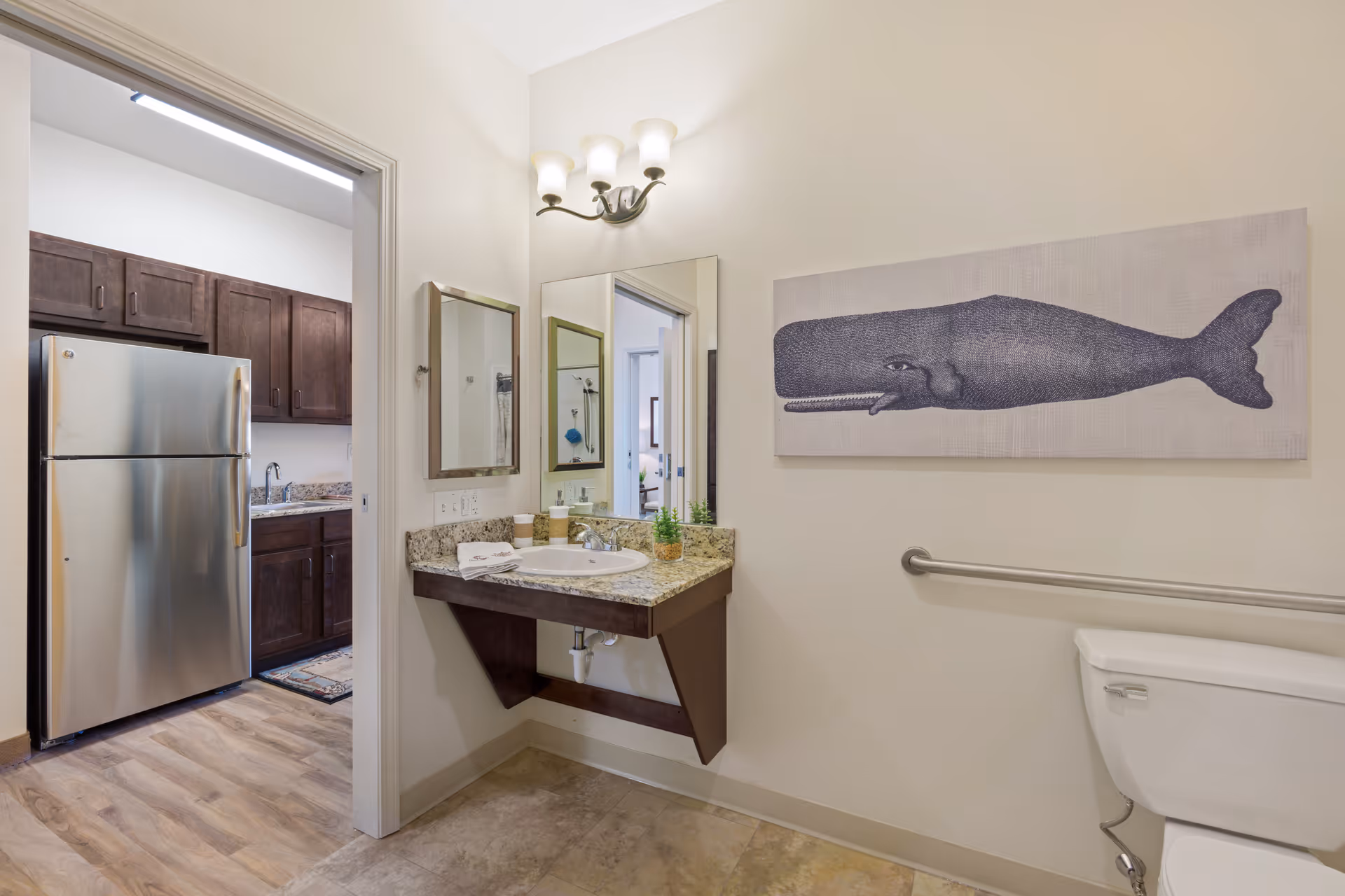 A bathroom with a wall-mounted sink featuring a granite countertop, a mirror above the sink, and a light fixture with three bulbs. To the right is a toilet with a grab bar mounted on the wall. Above the grab bar is a large artwork of a whale. Through the open door on the left, a kitchen area with a stainless steel refrigerator and dark wooden cabinets is visible.