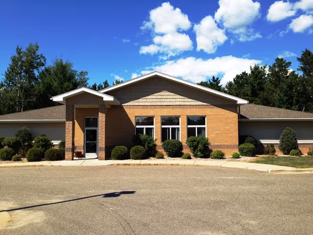 Single-story brick and siding building with a peaked roof, surrounded by small bushes and trees under a blue sky with scattered clouds.