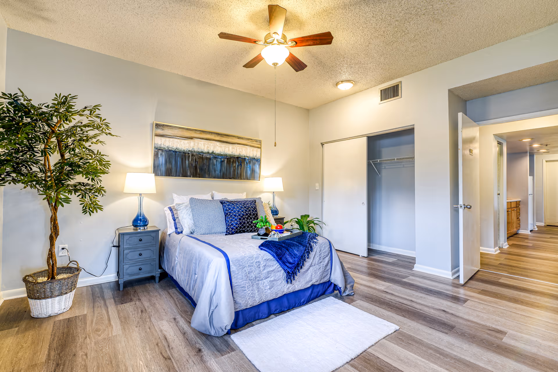 A neatly arranged bedroom with a bed dressed in gray and blue bedding, two bedside tables with lamps, a large potted plant, a ceiling fan with light, an open closet with sliding doors, and a hallway leading to another room.