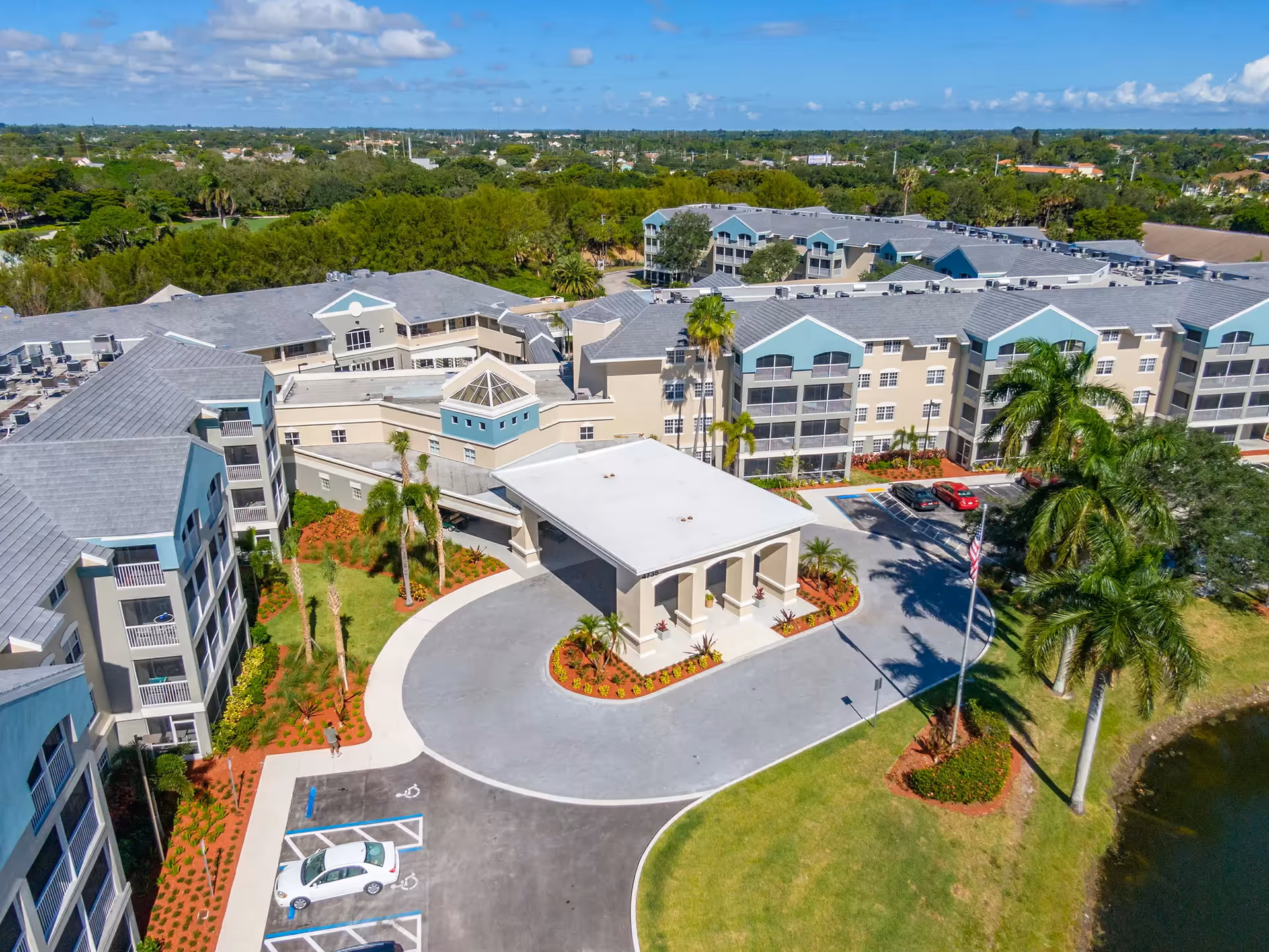 Aerial view of Discovery Village At Boynton Beach showing a large senior living facility with multiple connected buildings, a covered entrance driveway, landscaped grounds with palm trees, parking spaces including handicapped spots, and a small pond on the right side.