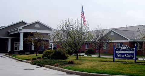 Front exterior view of Silver Oaks Health Campus building with a covered entrance, an American flag on a flagpole, small trees, and a sign displaying the facility name on a grassy lawn.