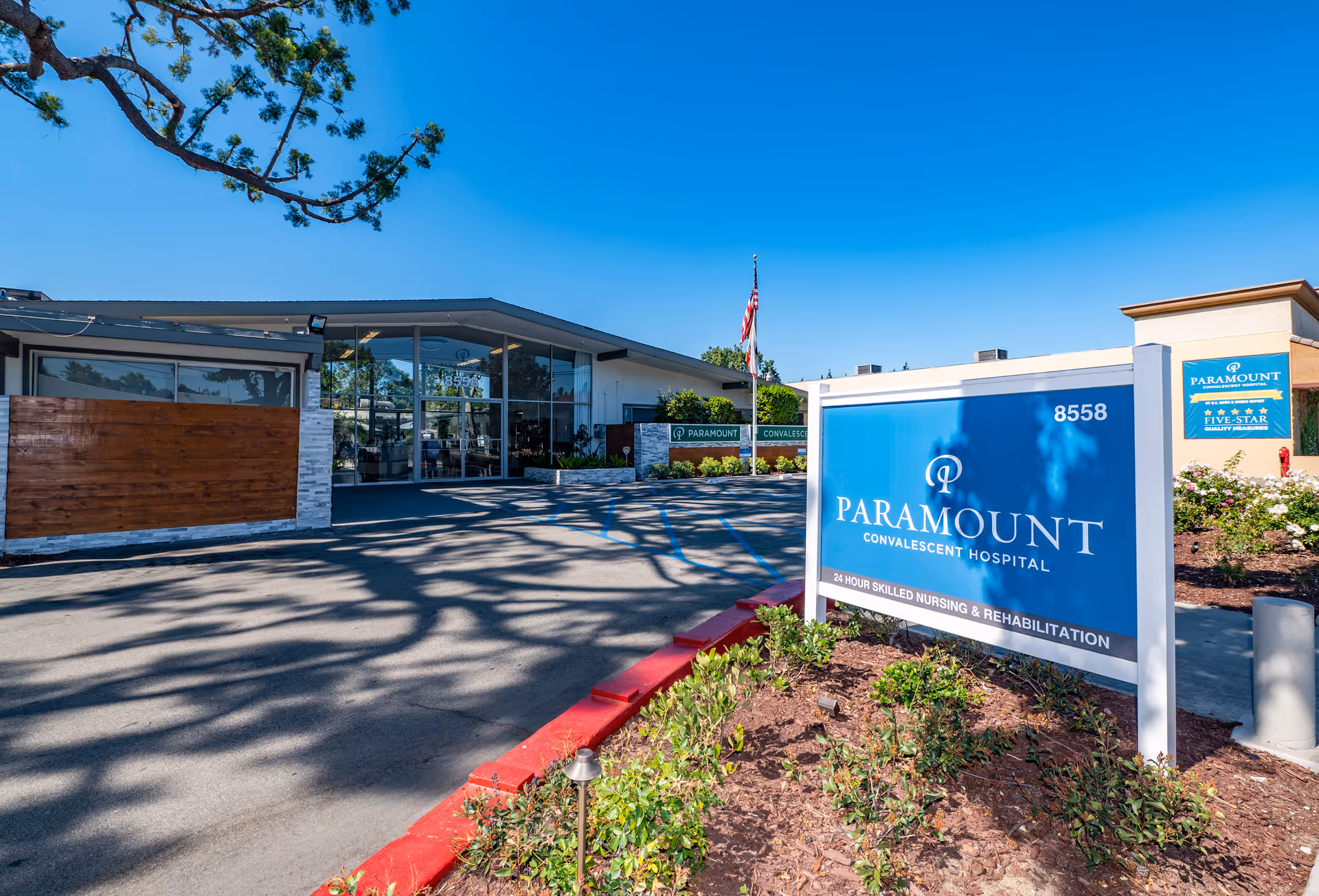 Exterior view of Paramount Convalescent Hospital showing the main entrance with large glass doors, a blue sign with the hospital name and address 8558, an American flag, and landscaped plants under a clear blue sky.
