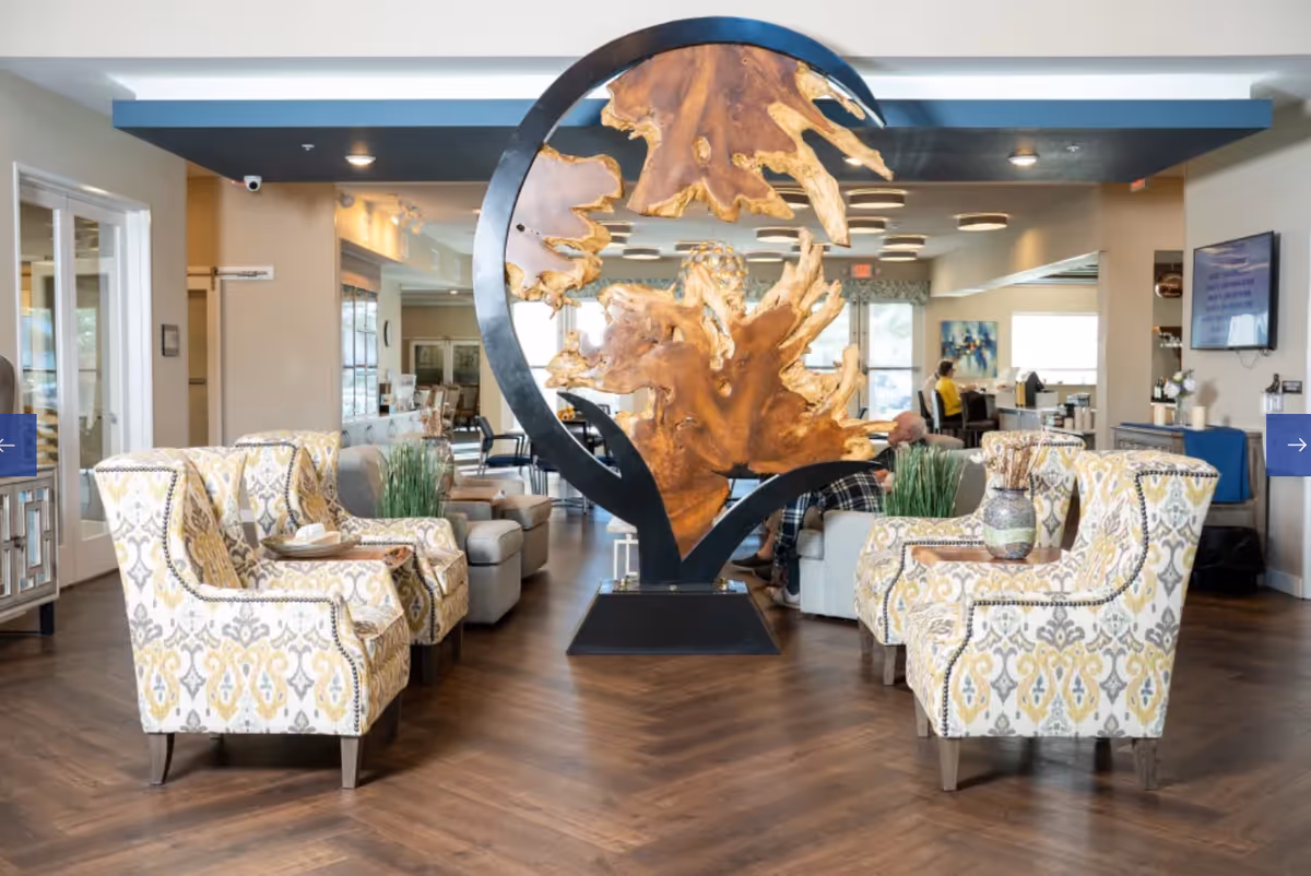 Bright communal lounge with patterned armchairs facing a large decorative wood-and-metal sculpture in the center.