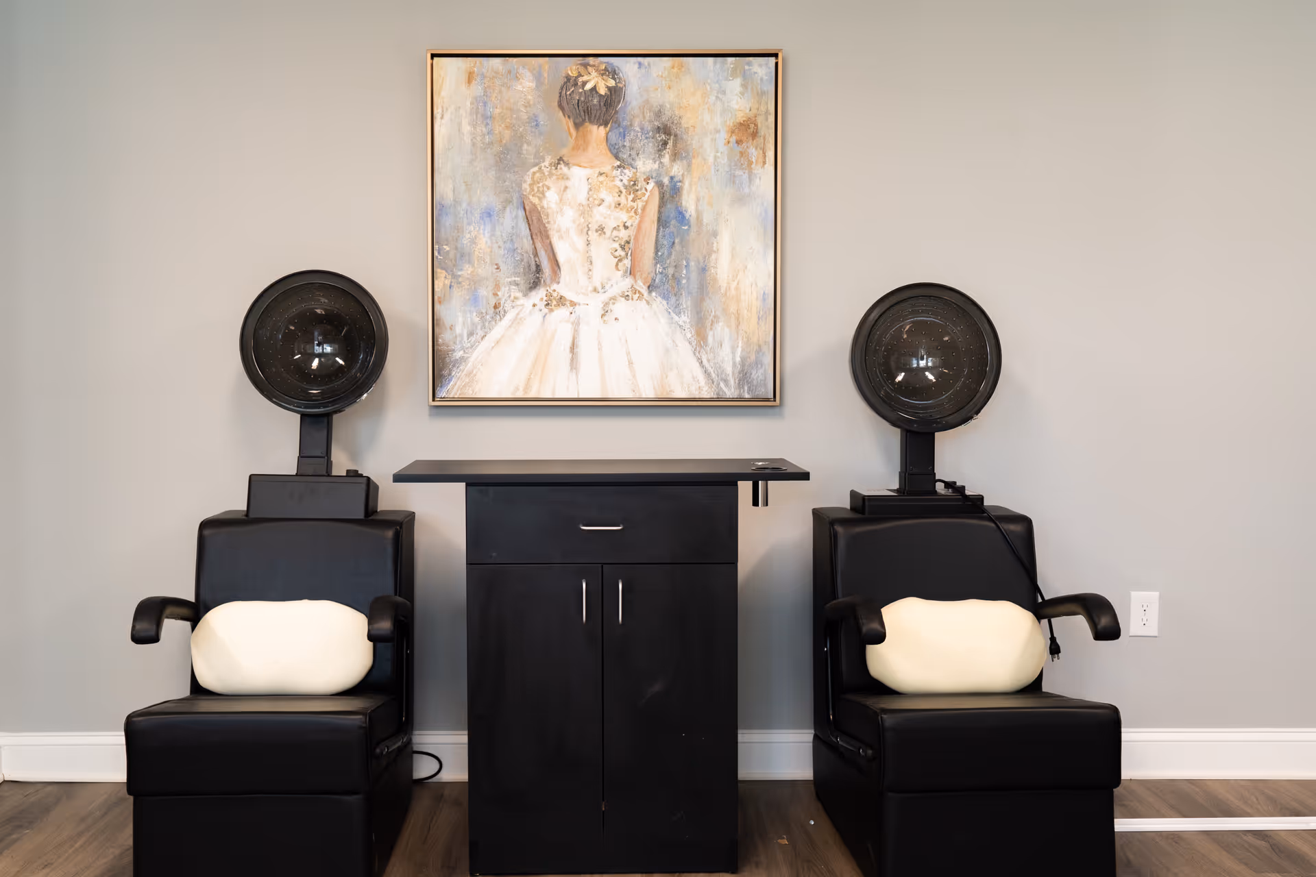 Two black salon hair dryer chairs with white cushions are positioned on either side of a black cabinet. Above the cabinet hangs a painting of a woman in a white dress seen from the back. The room has light gray walls and wooden flooring.