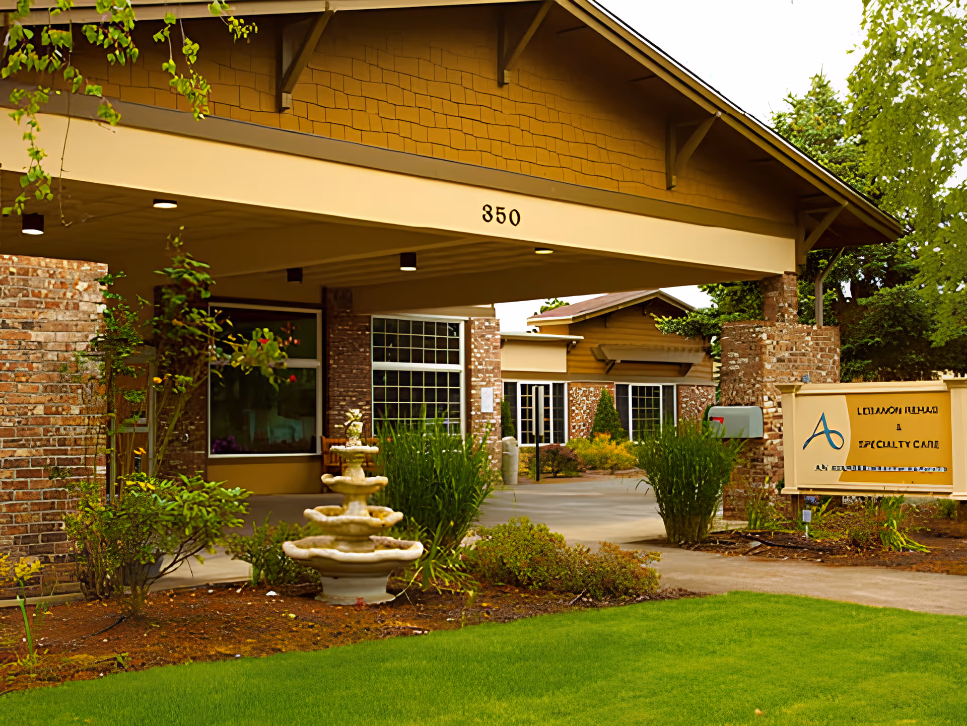 Entrance of a brick and wood building with a covered driveway, a three-tier stone fountain, green shrubs, and a sign indicating it is a rehabilitation and specialty care facility.
