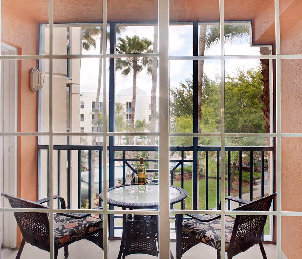 View through a window of a small balcony with two wicker chairs with floral cushions and a round glass-top table holding a bottle of white wine and two wine glasses. The balcony overlooks a garden area with palm trees and a building in the background.