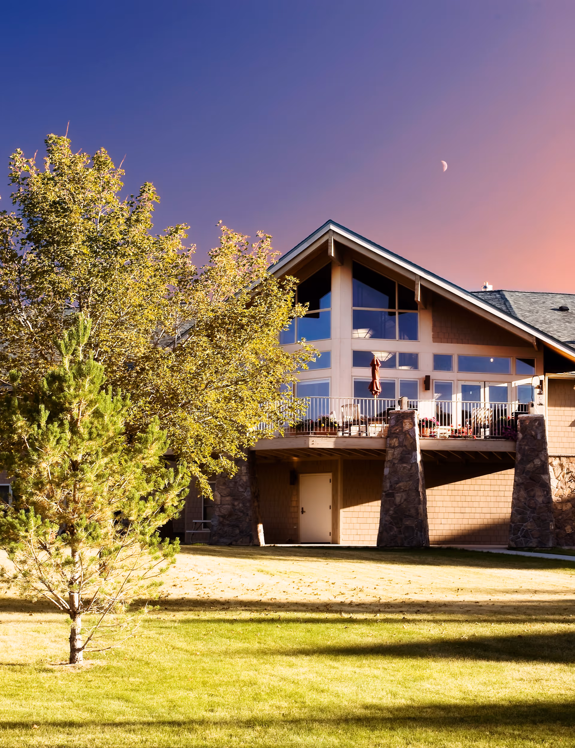 Exterior view of a senior living facility building with large windows and a balcony supported by stone pillars, surrounded by green grass and trees under a clear sky with a visible crescent moon.