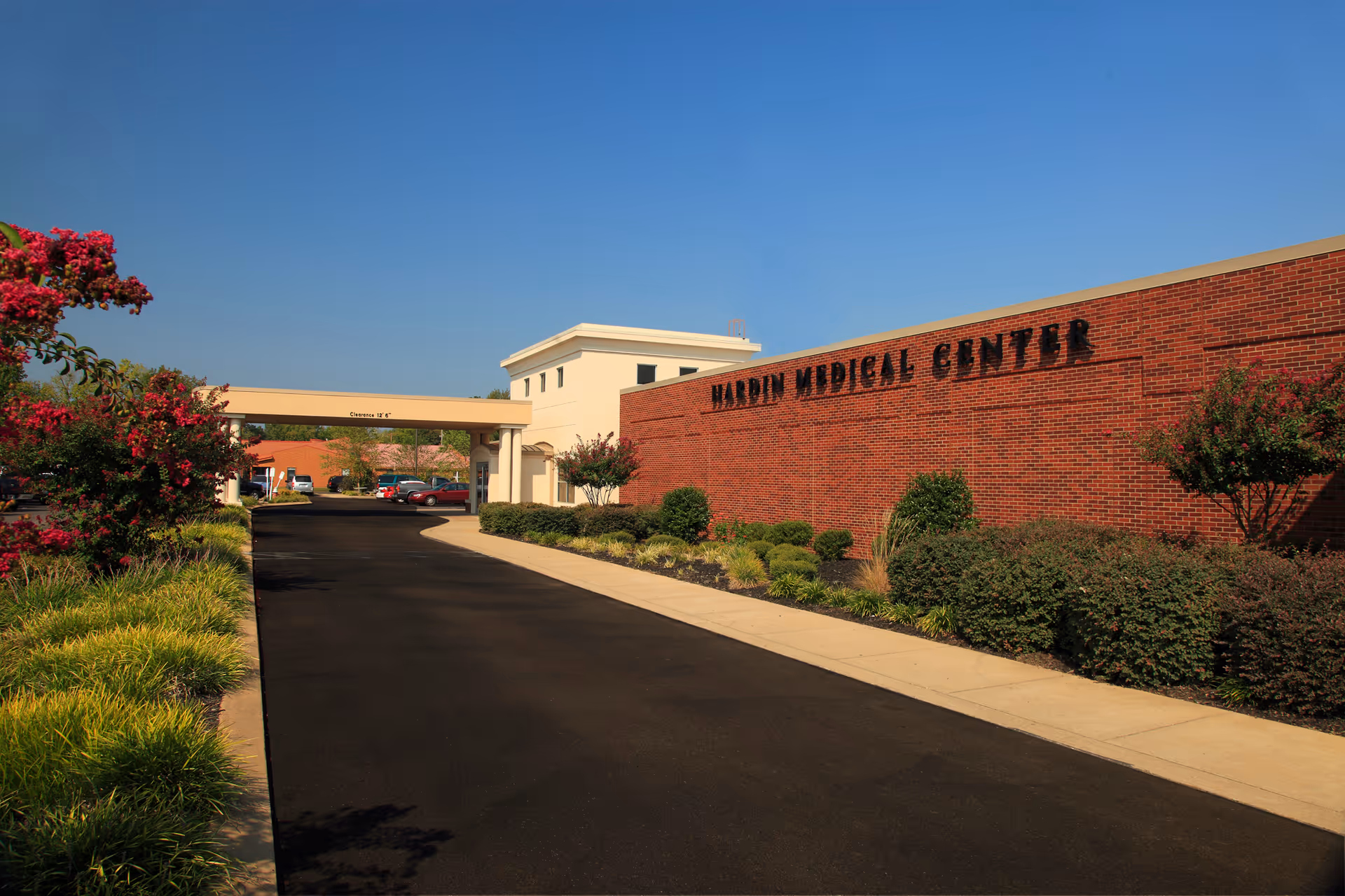 Driveway and entrance canopy leading to the brick facade of Hardin Medical Center with landscaped shrubs and a clear blue sky.