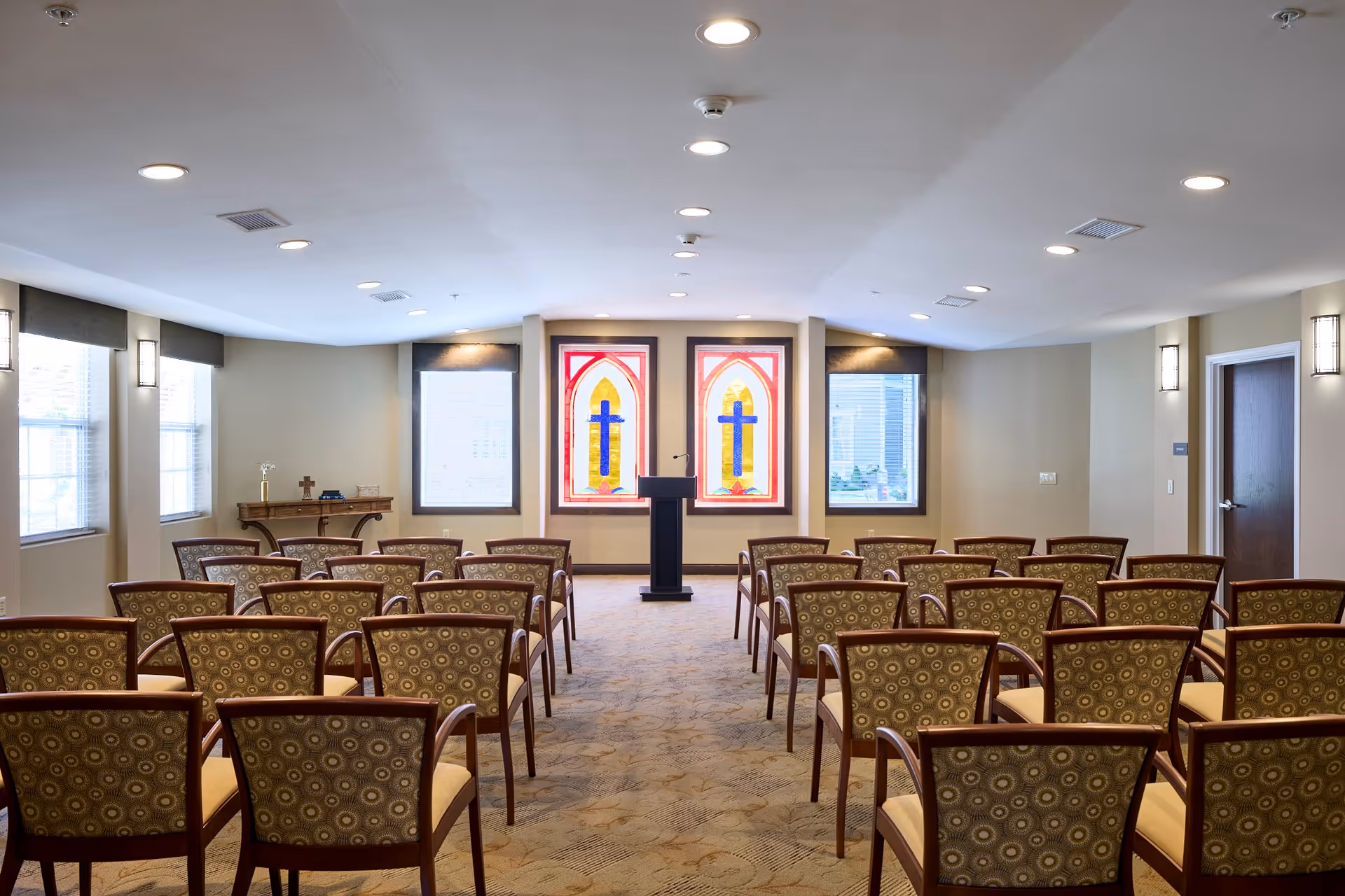 A small chapel or meeting room with rows of patterned chairs facing a podium. The room has beige walls, carpeted floor, and several windows, including two stained glass windows with blue crosses at the front.