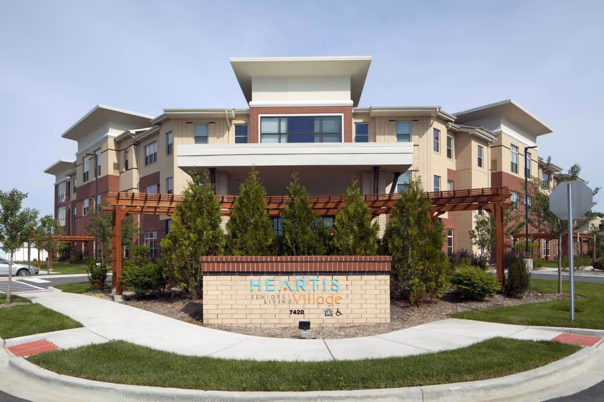 Exterior view of a multi-story senior living facility building with beige and red brick walls, large windows, and a covered entrance. In front of the building is a landscaped area with green shrubs and a brick sign that reads 'Heartis Senior Living Village 7420'. The sky is clear and the surrounding area includes sidewalks and a street.