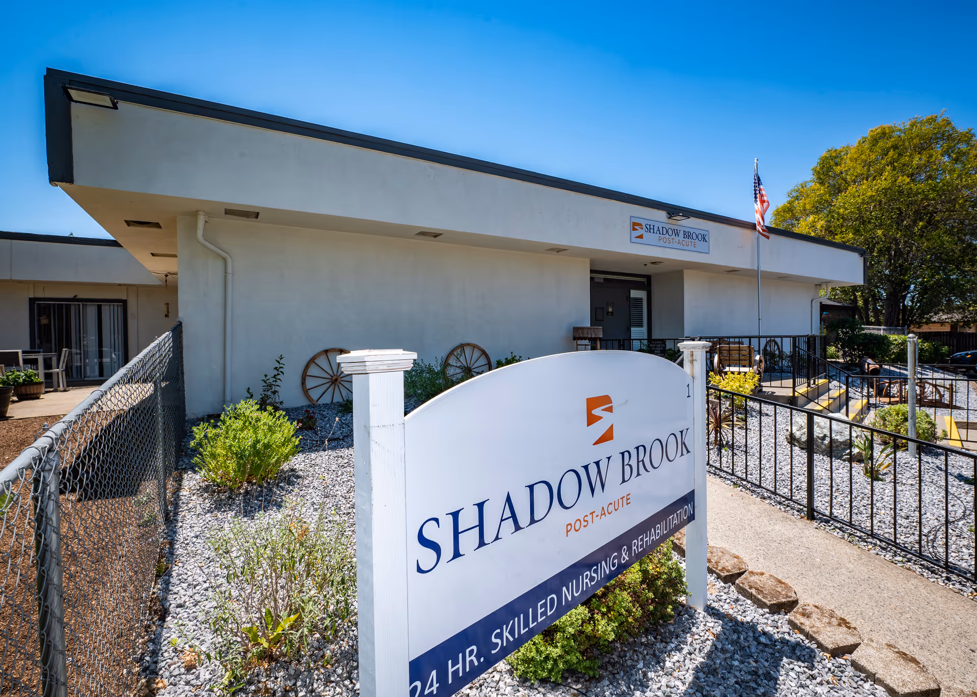 Exterior view of Shadow Brook Post-Acute facility showing the building entrance with a sign in the foreground that reads 'Shadow Brook Post-Acute 24 HR. Skilled Nursing & Rehabilitation'. There is a fenced garden area with plants and gravel, an American flag on a pole near the entrance, and clear blue sky above.