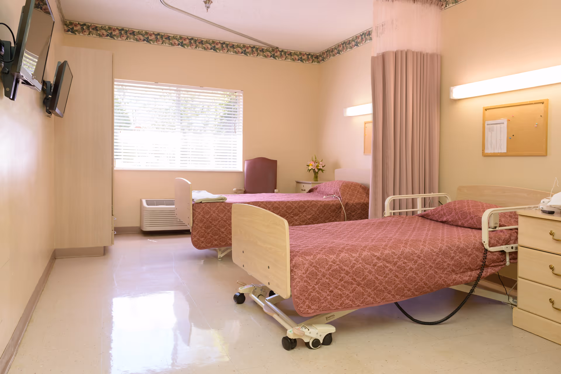 A bright room with two hospital-style beds covered in matching maroon patterned bedspreads. Each bed has a pillow and is equipped with side rails. There is a window with blinds letting in natural light, a maroon chair near the window, a small nightstand with flowers, and a curtain divider between the beds. The walls are light-colored with a floral border near the ceiling, and there are two wall-mounted lights and a cork bulletin board. A TV is mounted on the wall opposite the beds.