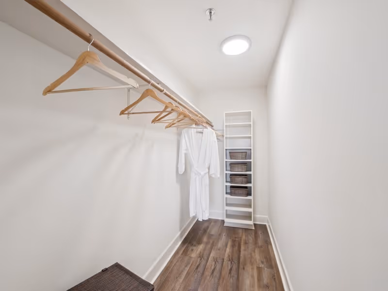 A walk-in closet with wooden hangers on a rod, a white robe hanging among them, a tall white shelving unit with woven baskets, and wood flooring under bright ceiling light.