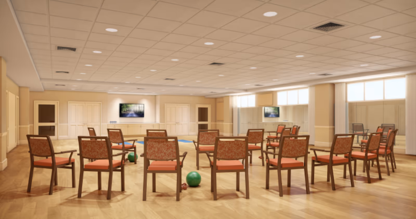 A spacious room with wooden flooring and a drop ceiling with recessed lighting. The room is arranged with multiple wooden chairs with red cushions placed in a circle. There are green exercise balls and small dumbbells on the floor, suggesting the room is used for group exercise or physical therapy. The walls are beige with white trim, and there are large windows letting in natural light. A flat-screen TV is mounted on the far wall above a cabinet.