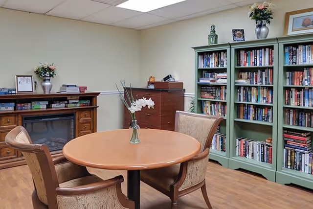 A cozy interior reading room with a round table and two chairs, bookshelves filled with books, and a decorative console.
