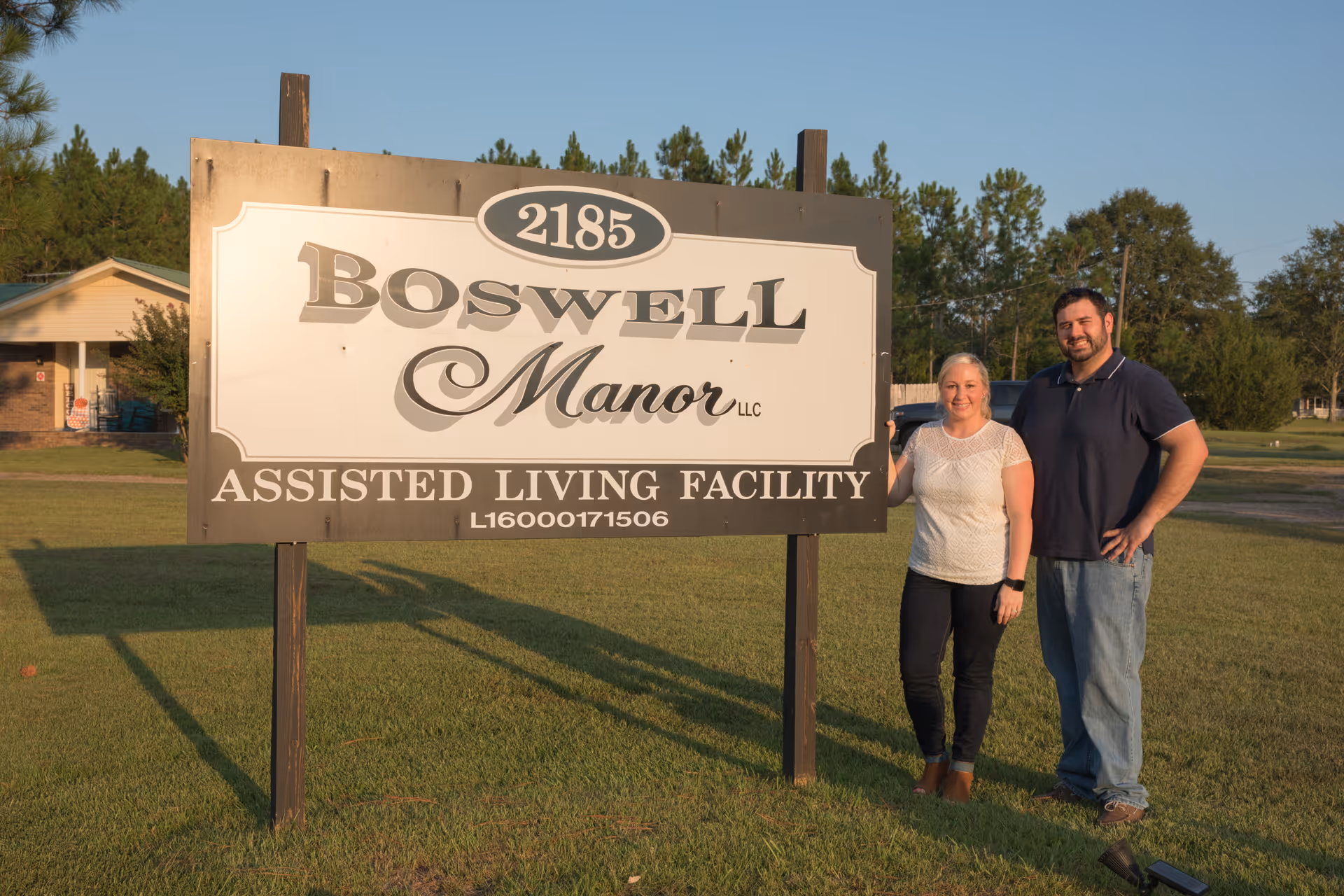 A man and a woman standing next to a large sign for Boswell Manor LLC Assisted Living Facility, located at 2185. The sign is set on a grassy area with trees and a building in the background under a clear sky.