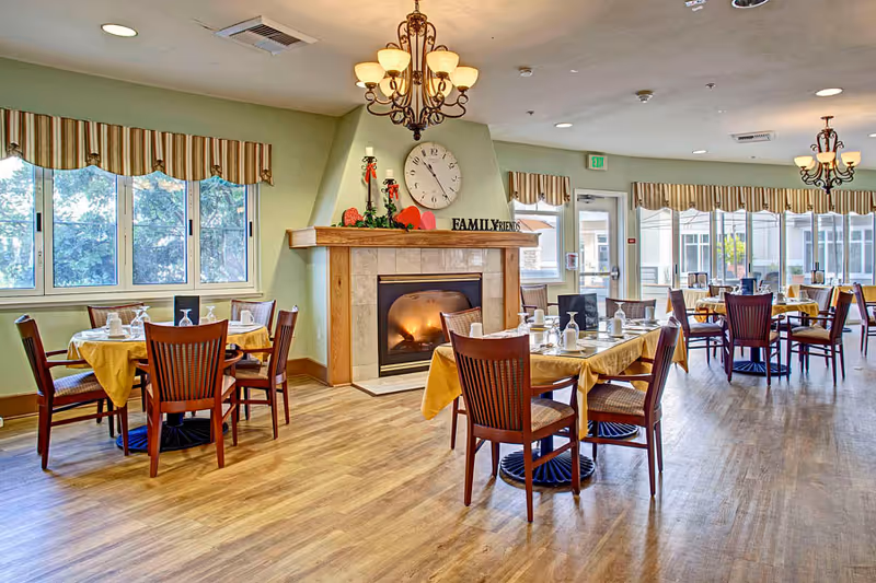 A bright dining room with several tables covered in yellow tablecloths, each set with white dishes and utensils. Wooden chairs surround the tables. A fireplace with a clock above it and decorative items including candles and a sign that says 'FAMILY FRIENDS' is visible. Large windows with striped valances let in natural light, and chandeliers hang from the ceiling.
