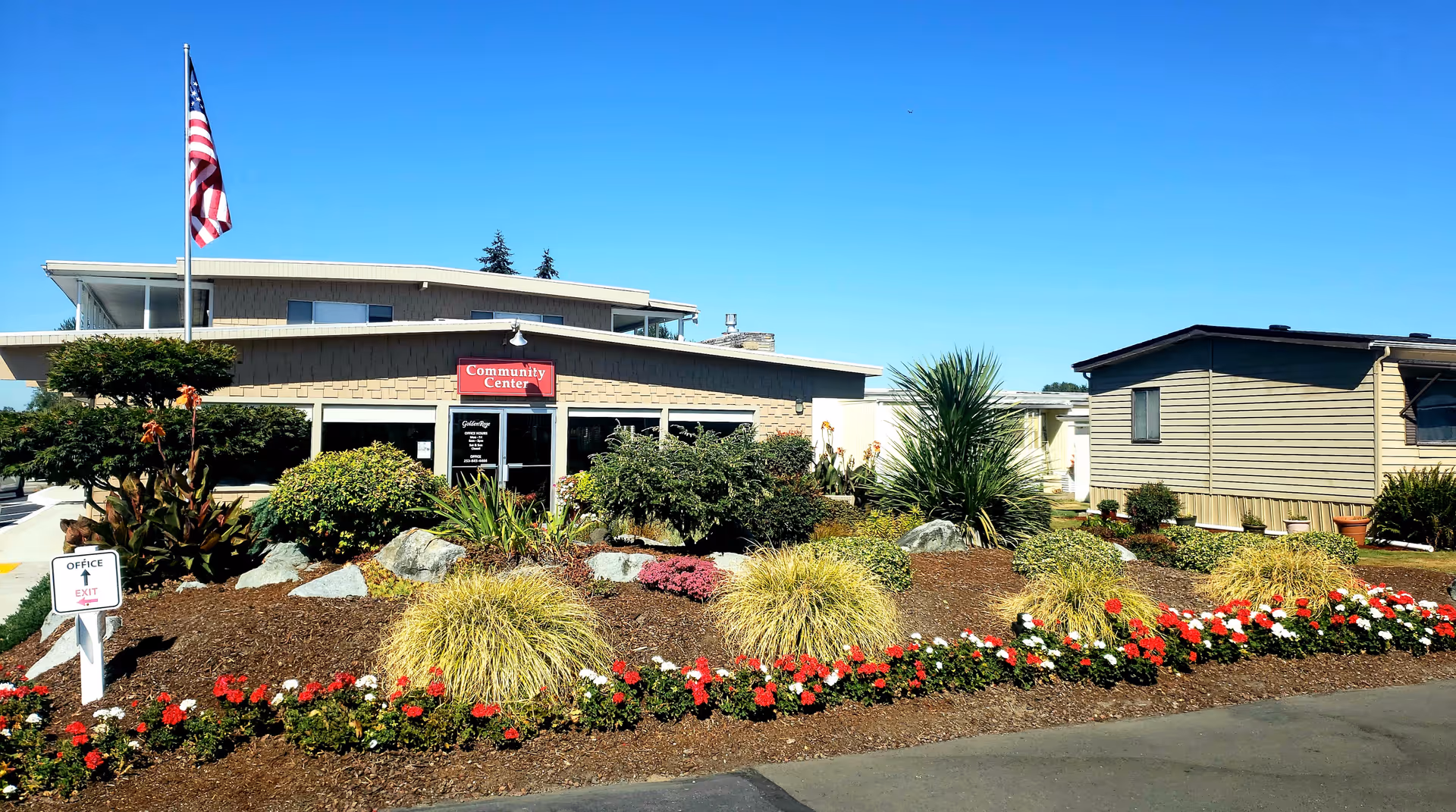 Exterior view of a community center building at Golden Rose 55+ Community near Tacoma, Washington. The building has a sign that reads 'Community Center' above the entrance. There is a well-maintained garden with various shrubs, flowers, and rocks in front of the building. An American flag is flying on a flagpole to the left of the building. A small sign near the garden indicates the office exit direction.