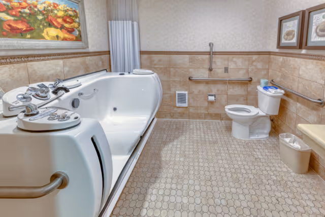 A spacious senior-friendly bathroom featuring a large walk-in bathtub with multiple control knobs and a handrail, a toilet with grab bars on the walls for support, beige tiled walls and floor, a small trash bin, and framed artwork on the walls.