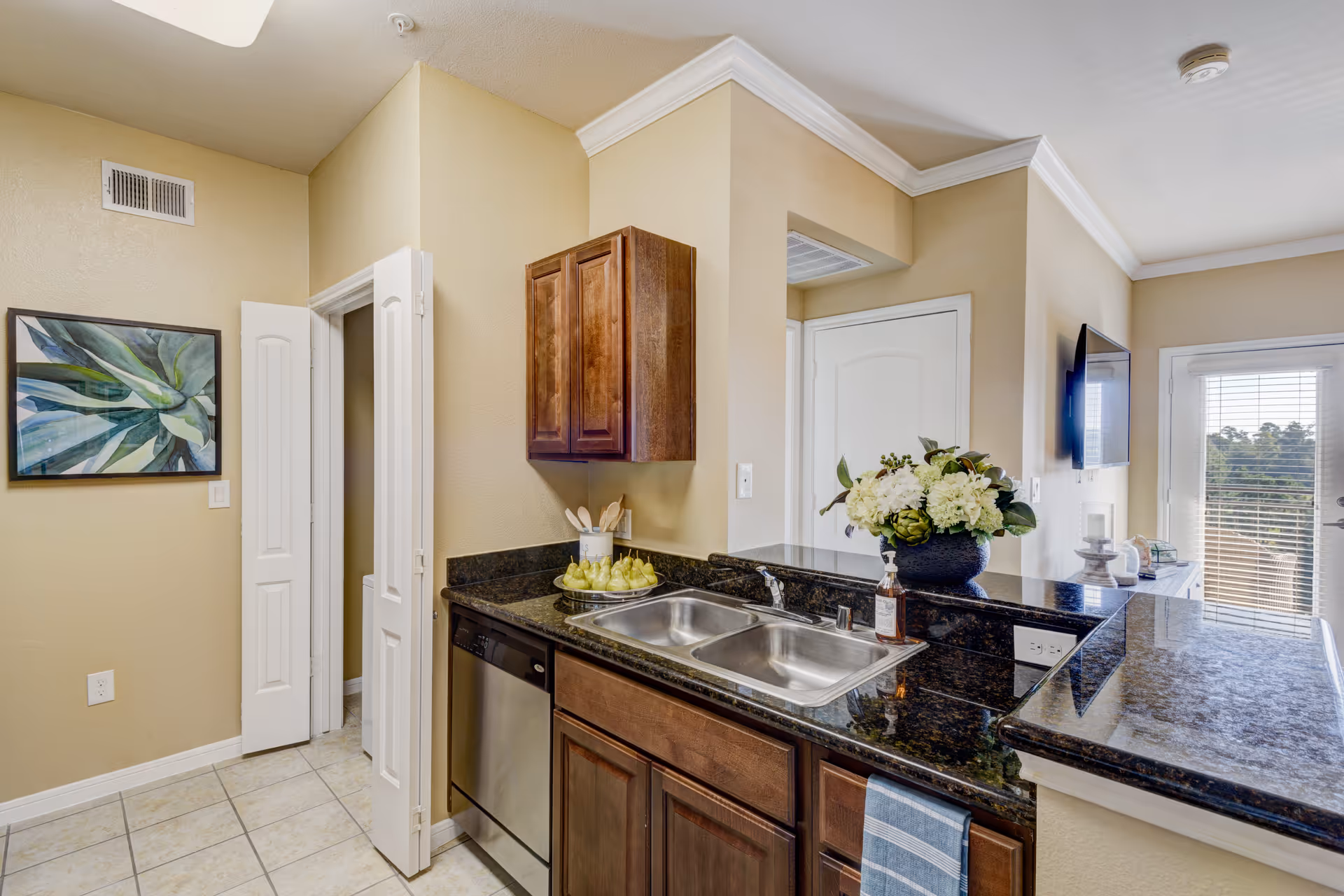 Open kitchen with granite countertops, stainless steel double sink and dishwasher, wooden cabinets, and a view into a bright living area.
