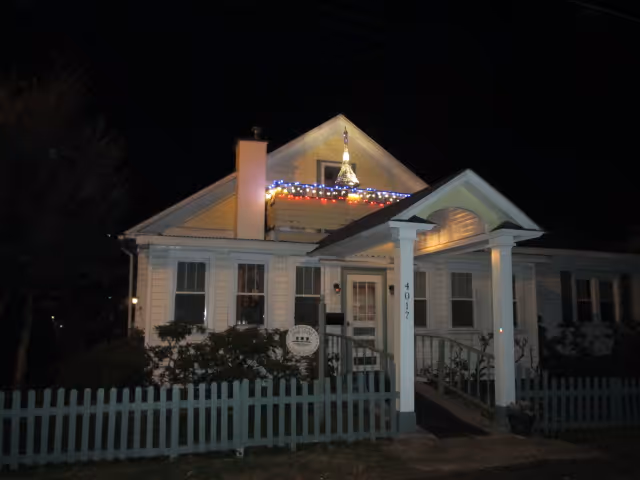 A white single-story house with a small porch and a picket fence in front, decorated with colorful string lights along the roofline, photographed at night.