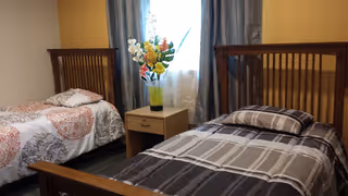 A bedroom with two single wooden beds, one with a patterned white and red bedspread and the other with a gray and white striped bedspread. Between the beds is a wooden nightstand with a vase of colorful flowers. A window with sheer white curtains and gray drapes is behind the nightstand.