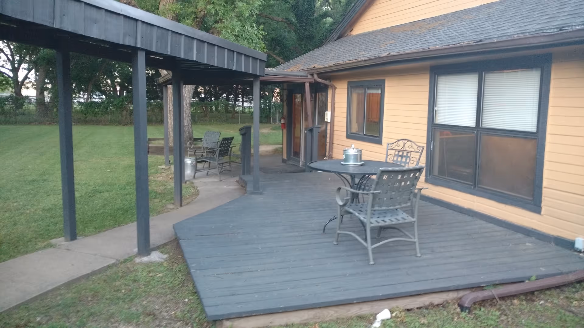Outdoor patio area at Brixton Village Assisted Living with a wooden deck, metal table and chairs, a covered walkway, and a yellow building with windows in the background surrounded by grass and trees.