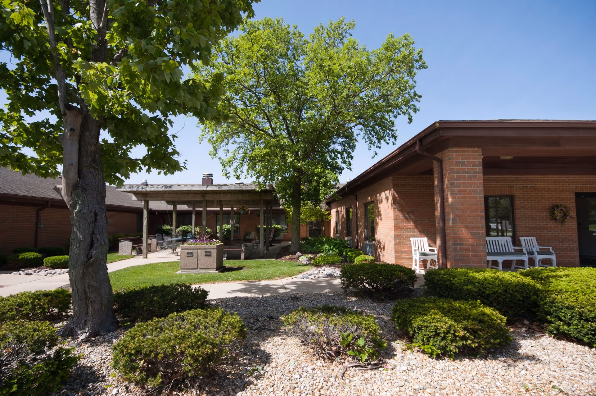 Outdoor courtyard area at Accolade Healthcare of Paxton on Pells featuring a brick building with white chairs on the porch, a paved walkway, green bushes, trees, and a covered seating area with patio furniture under a wooden pergola.