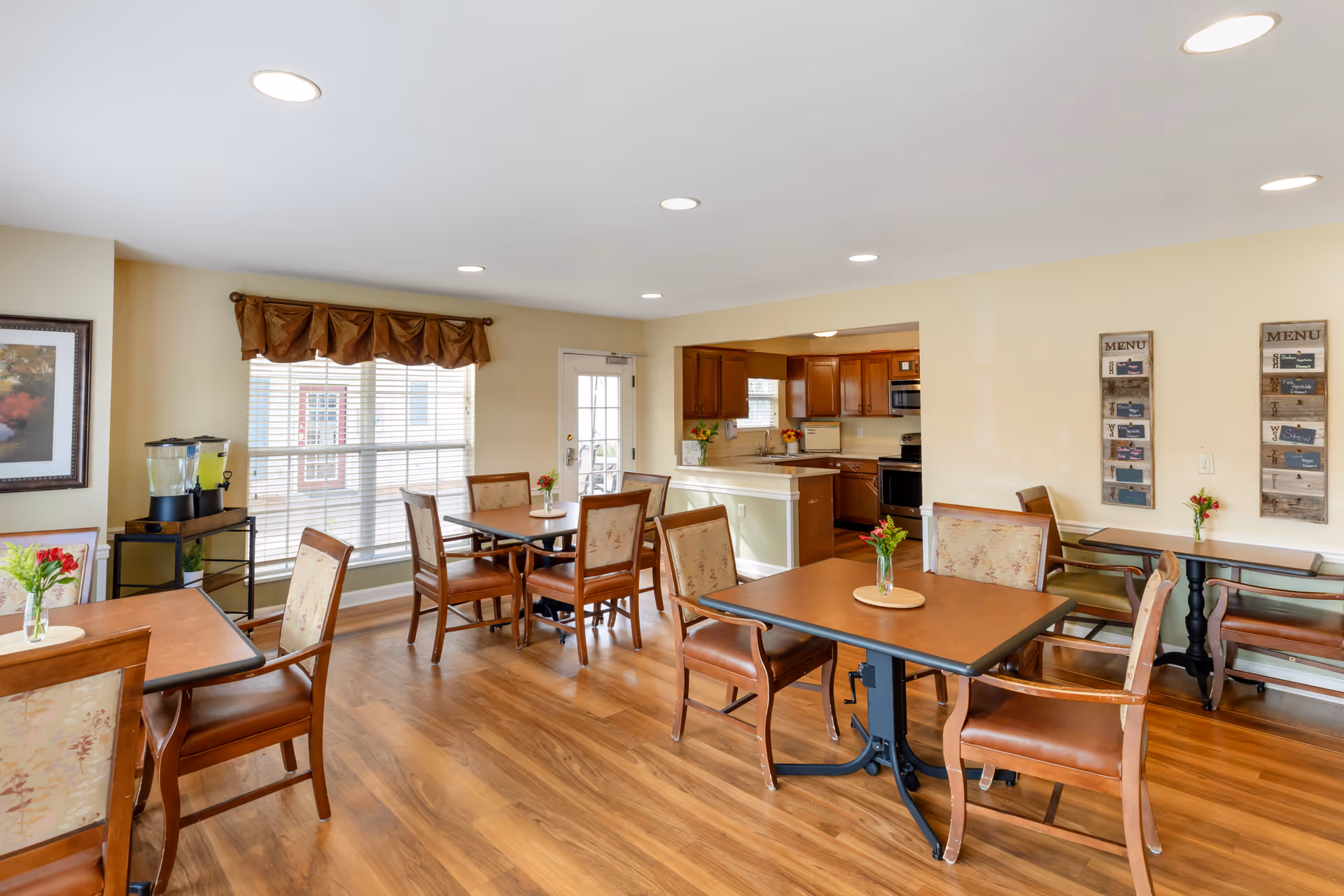 A bright dining area with wooden floors and several tables surrounded by cushioned chairs. Each table has a small vase with flowers. The room has large windows with blinds and a brown valance. In the background, there is a kitchen with wooden cabinets and stainless steel appliances. Two menu boards are mounted on the wall to the right.