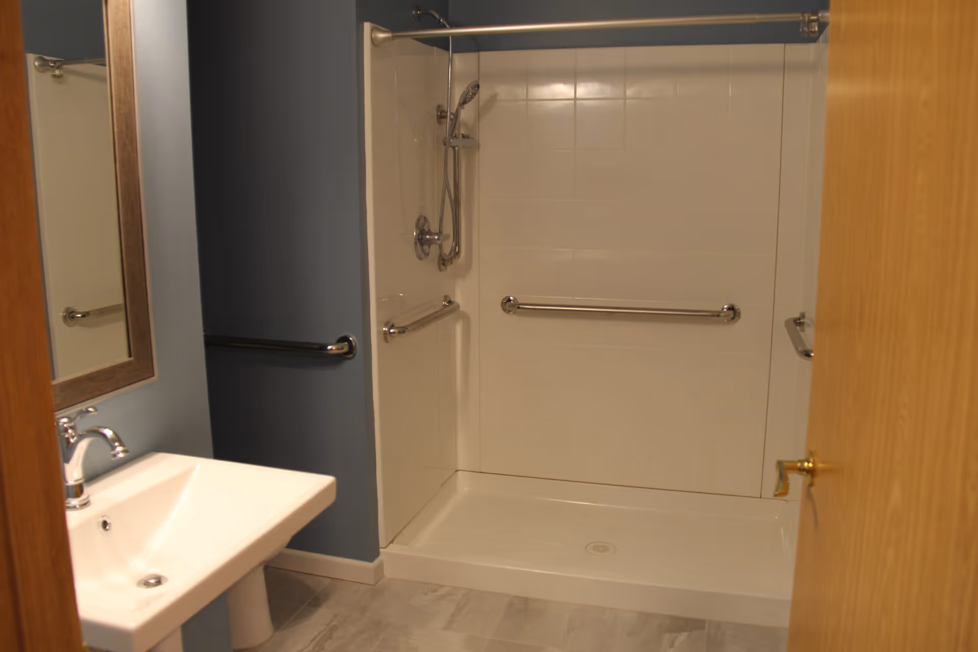 A bathroom with a walk-in shower featuring white tiled walls and multiple stainless steel grab bars. There is a handheld showerhead attached to the wall. A white pedestal sink with a chrome faucet is visible on the left side, along with a mirror above it. The walls are painted blue, and the floor has light-colored tiles. A wooden door is partially open on the right side.
