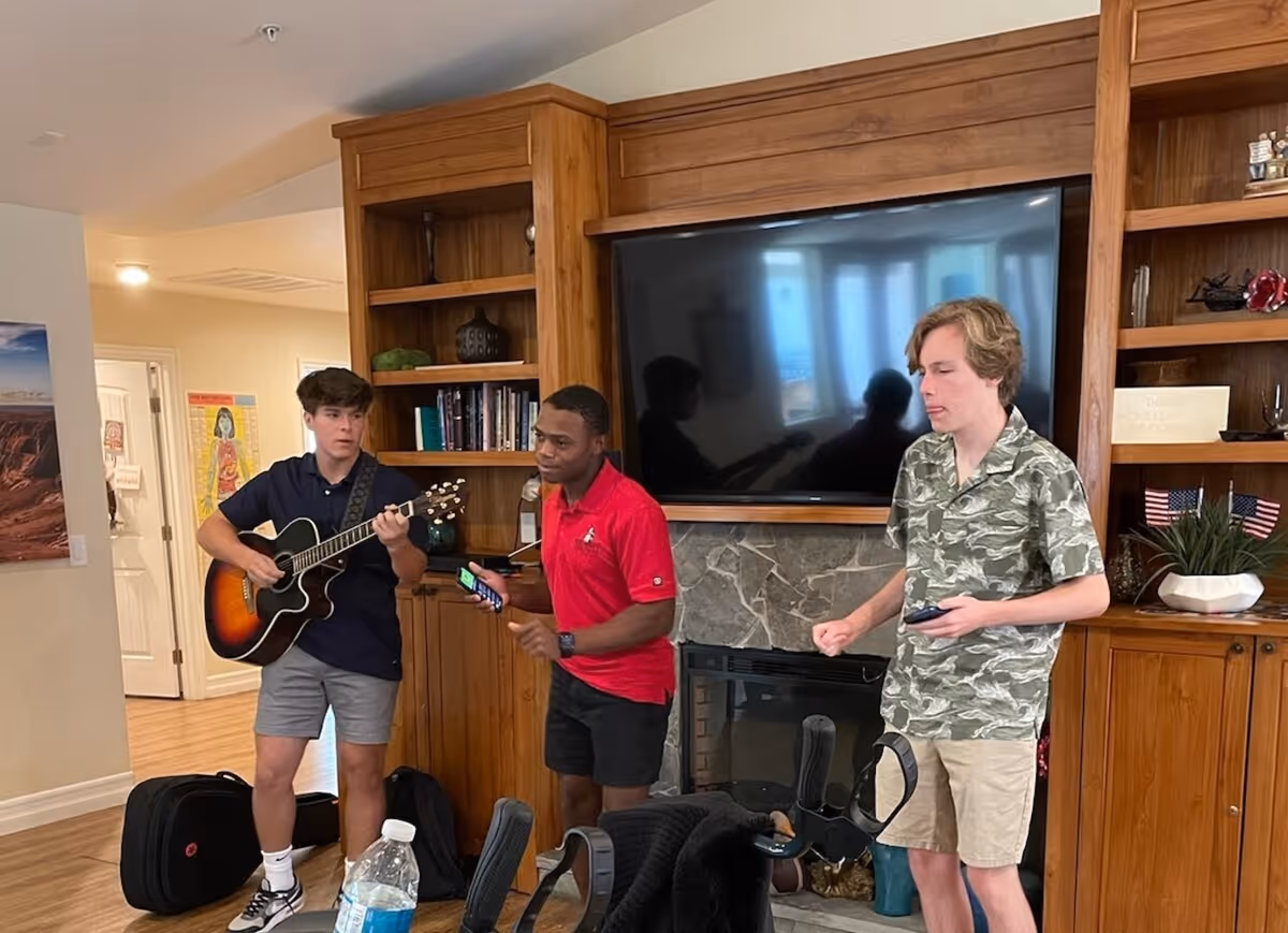 Three young men performing music in a living-room style common area, one playing an acoustic guitar in front of a TV and fireplace.
