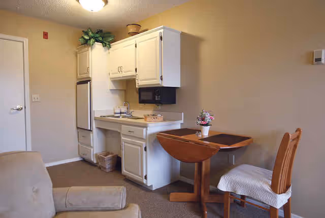 A small kitchenette area with white cabinets, a sink, a microwave, and a mini refrigerator. Next to the kitchenette is a wooden drop-leaf table with a flower pot on top and a wooden chair with a white cushion. Part of a beige armchair is visible in the foreground. The walls are painted beige and the floor is carpeted.