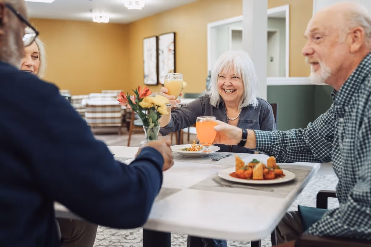 Four elderly people sitting around a dining table in a senior living facility, smiling and raising their glasses in a toast. The table has plates of food and a vase with flowers. The room has warm lighting and framed artwork on the walls.