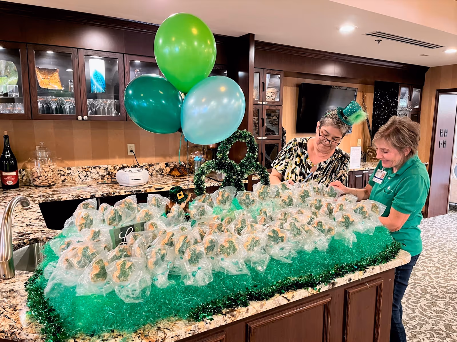Two women standing behind a kitchen island decorated with green tinsel and balloons, arranging individually wrapped treats. The kitchen has granite countertops, dark wood cabinets with glass doors, and various kitchen items including a large bottle and a jar of corks.