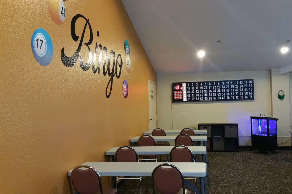 Interior room with several tables and chairs arranged in rows facing a bingo number board on the wall. The wall on the left side is decorated with the word 'Bingo' and colorful bingo balls with numbers. There is a small cabinet and a blue-lit aquarium in the corner of the room.