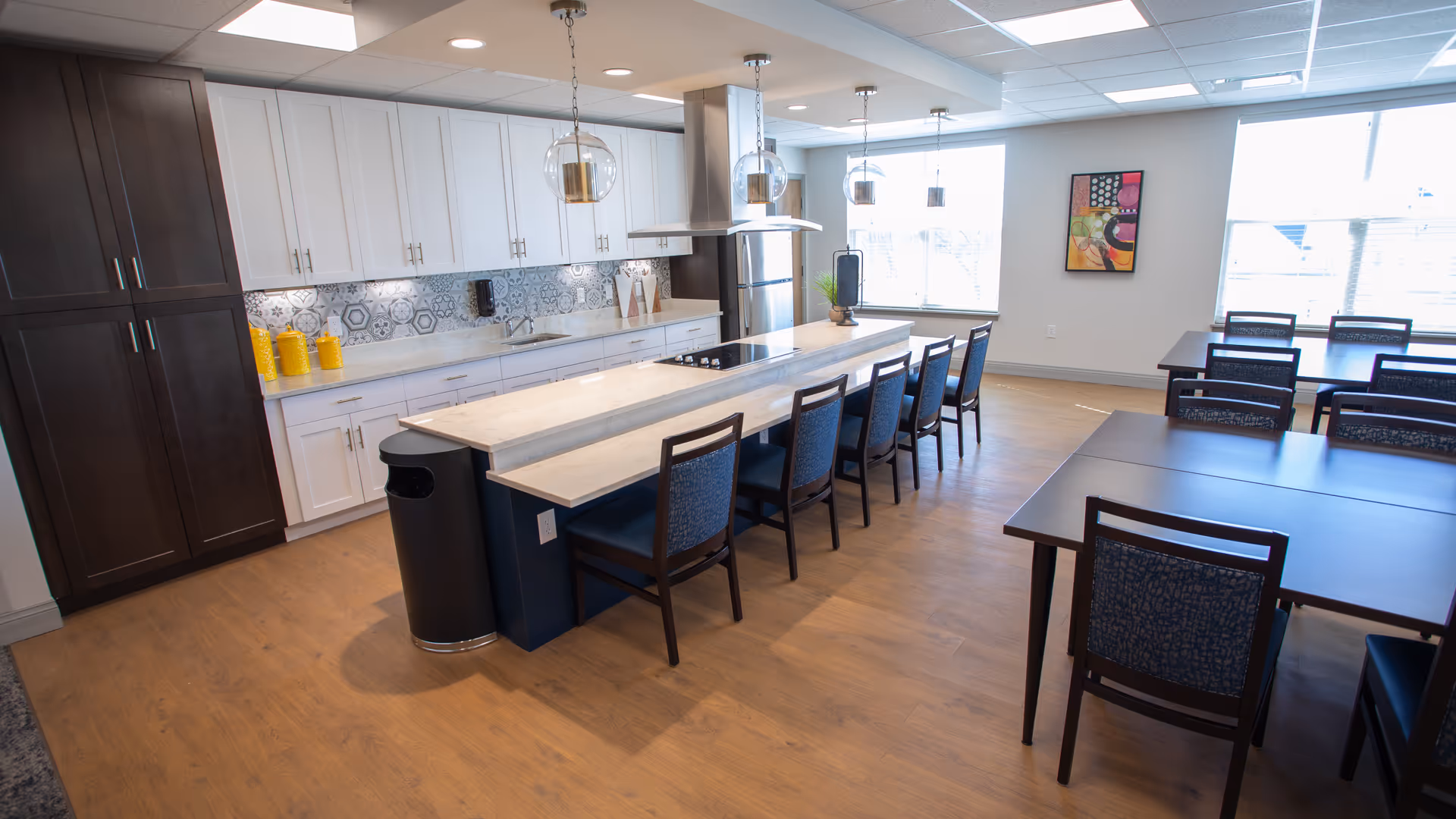 A bright and modern dining area with a long kitchen island featuring a marble countertop and several chairs lined up along it. The kitchen has white upper cabinets, dark lower cabinets, a stainless steel refrigerator, and a decorative backsplash. There are pendant lights hanging above the island, and additional tables and chairs are arranged near large windows letting in natural light. A colorful abstract painting is mounted on the wall.