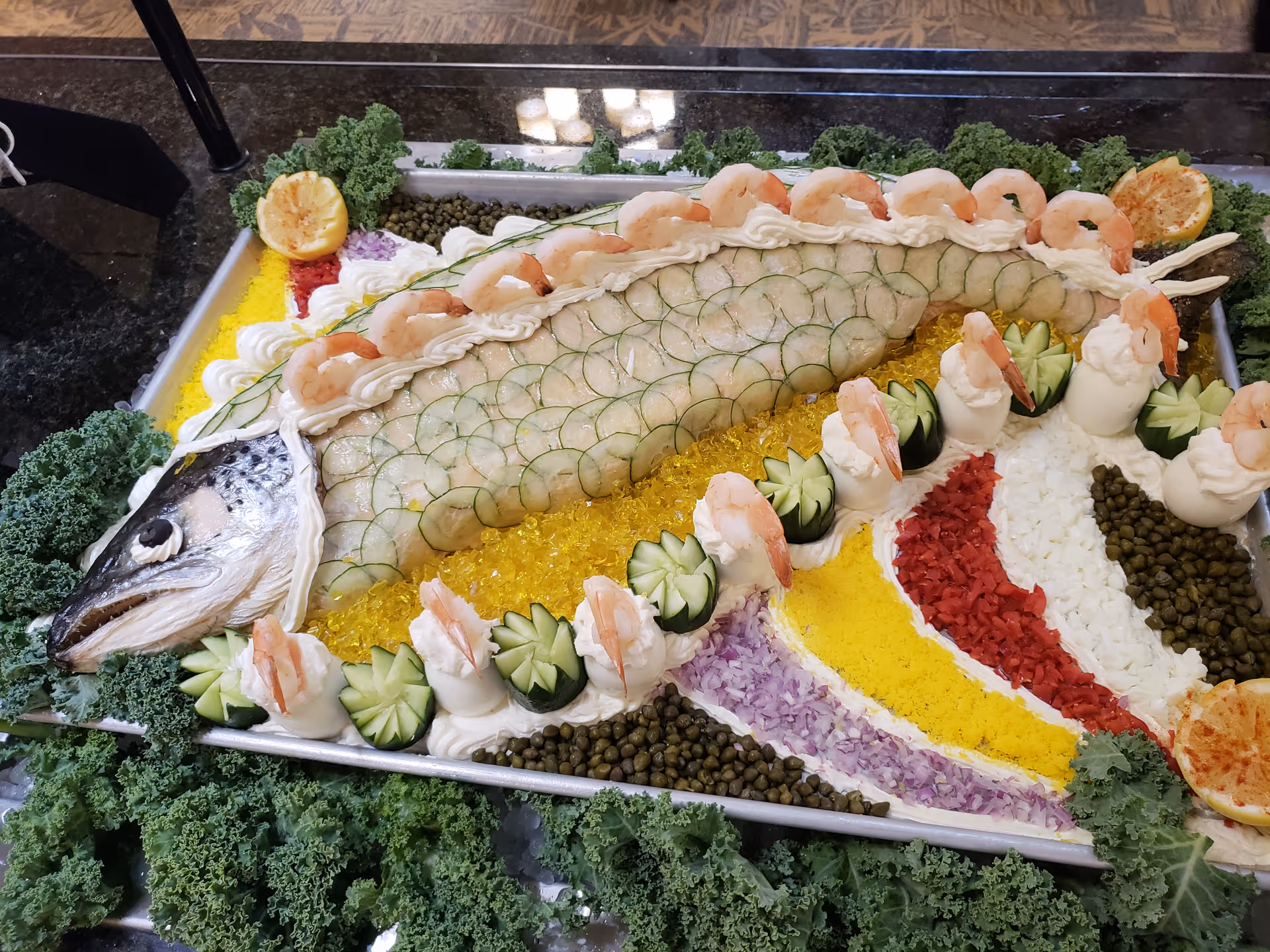 A large decorative seafood platter featuring a whole fish with its body covered in thin cucumber slices and topped with shrimp. The platter is garnished with various colorful chopped vegetables, capers, lemon halves, and leafy greens surrounding the tray.