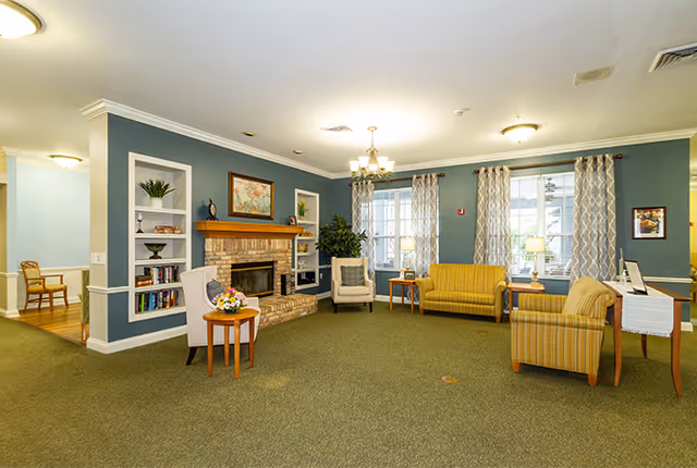 A cozy living room area in an assisted living facility featuring a brick fireplace with a wooden mantle, built-in shelves with decorative items, a chandelier, and multiple seating options including armchairs and a sofa. The walls are painted blue with white trim, and large windows with patterned curtains allow natural light to enter the room.