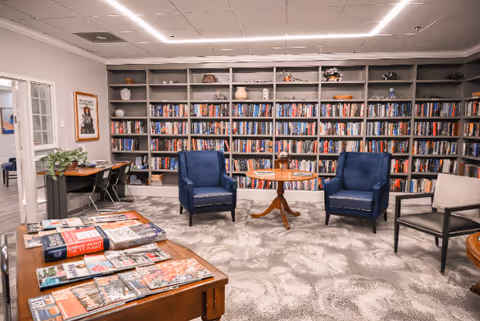 A cozy library room with a large bookshelf filled with books along the back wall. In front of the bookshelf are two blue armchairs with a small round wooden table between them. To the left, there is a wooden table with magazines and books on it, and a small desk with a plant and chairs. The room has a light-colored patterned carpet and bright overhead lighting.