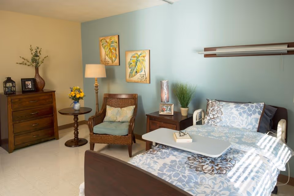 A bright, homey patient room featuring a hospital bed with floral bedding, a bedside table, armchair, dresser and decorative accents.
