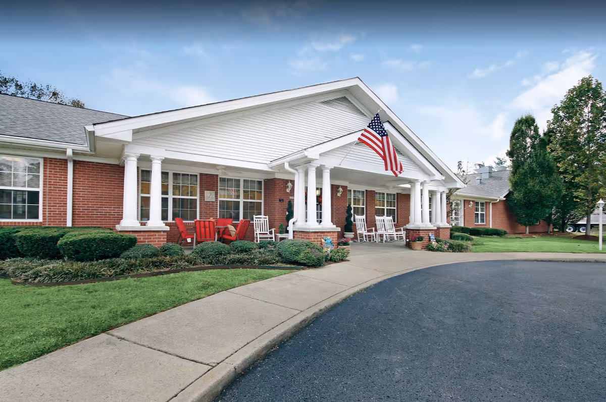 Brick single-story senior living facility entrance with white columns, rocking chairs, landscaping, and an American flag.