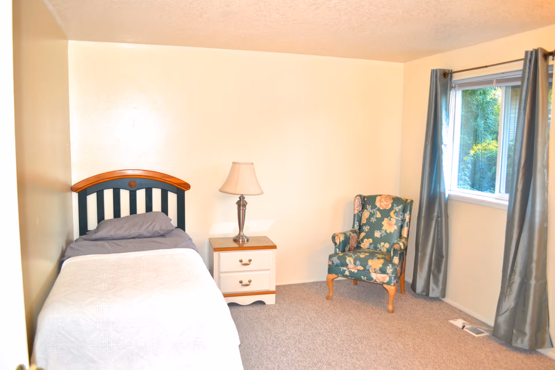 A simple bedroom with a single bed featuring a wooden headboard, a bedside table with a lamp, a floral upholstered armchair, and a window with gray curtains letting in natural light.