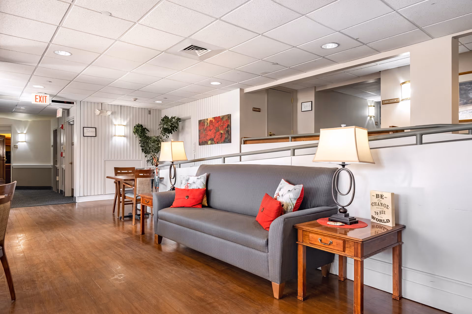 A cozy senior living facility common area with a gray sofa adorned with red and floral pillows, two wooden side tables each with a lamp, a wooden dining table with chairs, a potted plant, and wall art featuring red flowers. The space has wood flooring, white paneled walls, and a ceiling with recessed lighting and an exit sign visible in the background.