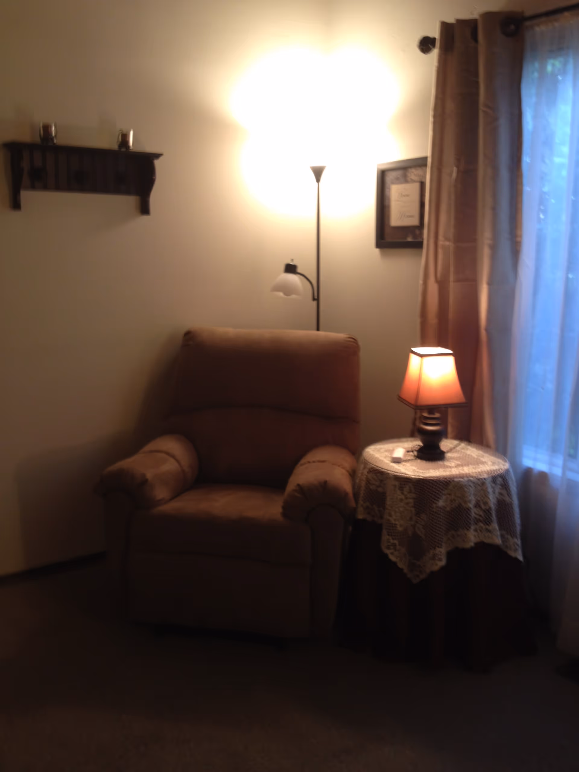 A cozy corner of a room featuring a brown upholstered armchair next to a round side table covered with a lace tablecloth. On the table is a small lamp with a warm orange shade. Behind the chair is a tall floor lamp turned on, casting light on the wall. A framed picture hangs on the wall next to the window with beige curtains.