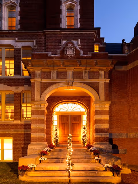 Entrance of a brick building at dusk with warm lights glowing from the windows. The entrance features a large wooden double door framed by an ornate stone archway. Stone steps lead up to the door, decorated with small potted plants and string lights wrapped around the handrails.