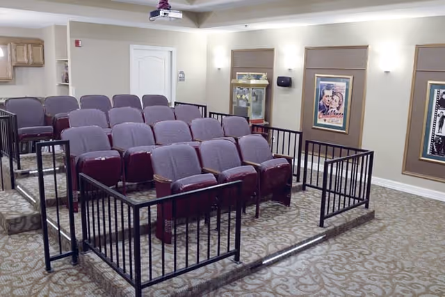 A small theater room with three rows of purple cushioned seats arranged on a carpeted floor. The room has beige walls with framed movie posters and a display cabinet in the corner. There is a projector mounted on the ceiling.