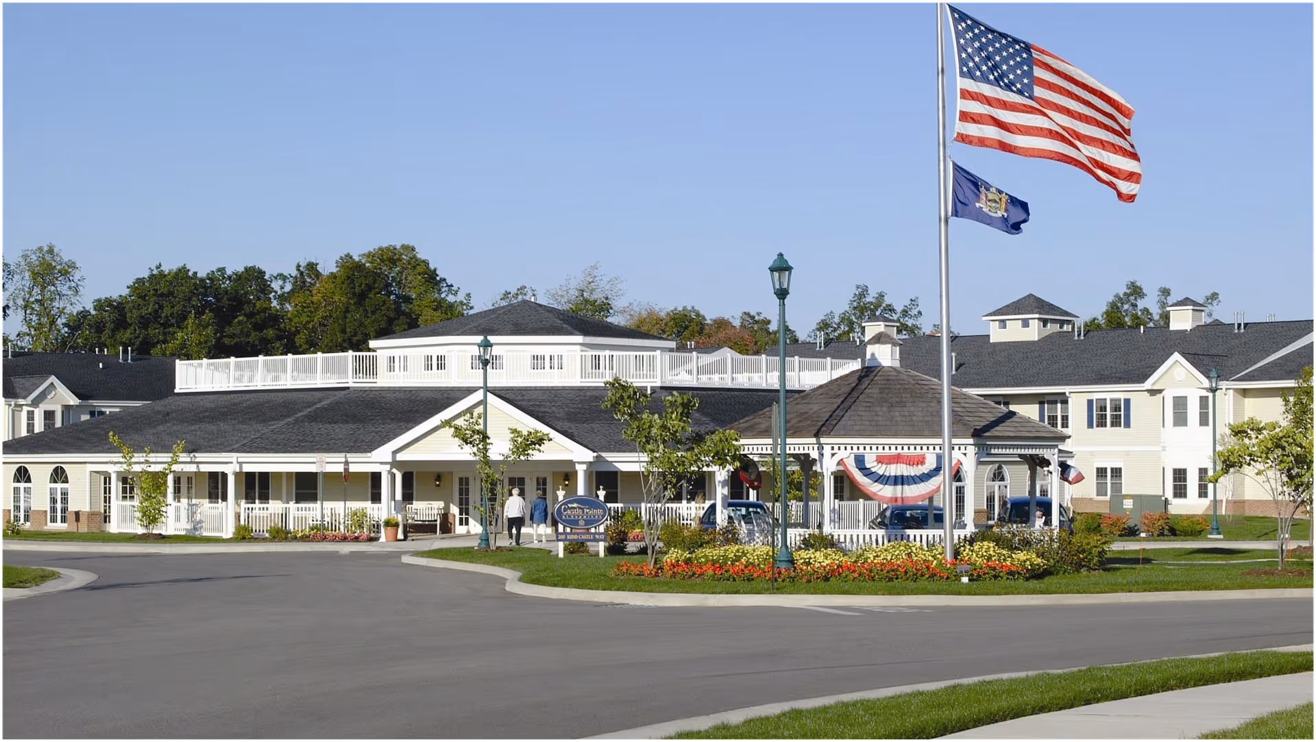 Exterior view of Castle Pointe at Baywinde senior living facility showing a large building with white and beige siding, a covered entrance, a gazebo decorated with patriotic bunting, two flagpoles with the American flag and a state flag, and landscaped grounds with flowers and trees under a clear blue sky.